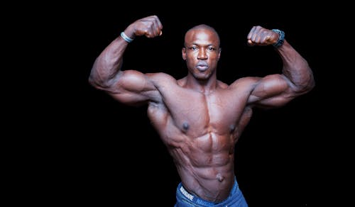 Muscular shirtless African American man standing with raised arms and looking at camera while flexing muscles during training on black background
