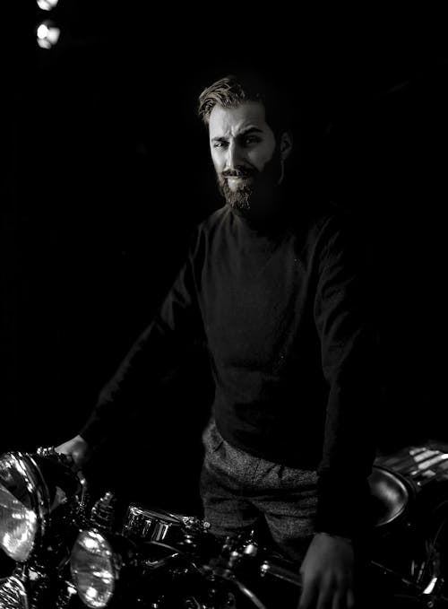 Black and white male in turtleneck standing with motorcycle on black background and looking at camera