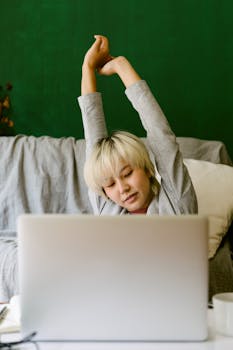Asian woman stretching arms while using laptop in a cozy living room setting.