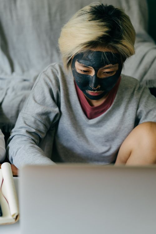 Free Serious young Asian female in casual wear with black facial mask working on netbook and looking at screen while sitting at comfortable sofa Stock Photo