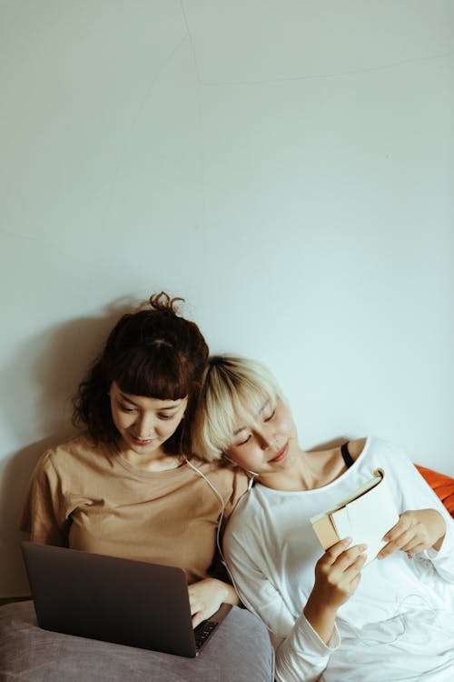 Positive young Asian sisters wearing casual clothes using modern netbook and reading book while lying on floor beside each other against gray wall