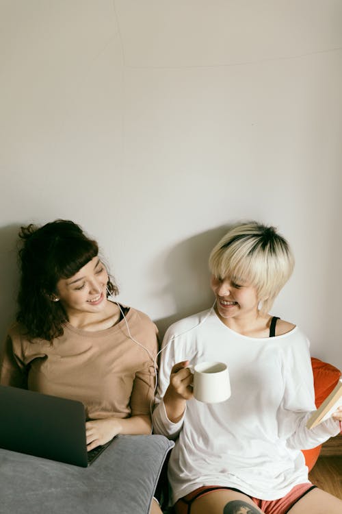 Free From above of smiling young Asian girlfriends in casual clothes sitting on floor with cup of hot beverage and browsing netbook while looking at each other Stock Photo