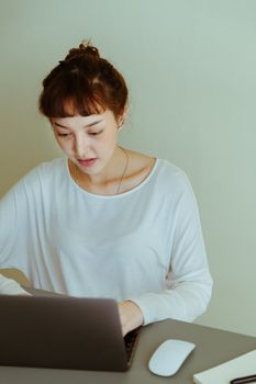 Serious young Asian female in white casual clothing using modern laptop and sitting at table at home against beige wall