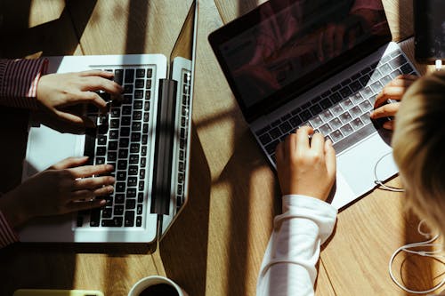 Free From above crop anonymous females browsing netbooks while sitting at wooden table with cup of coffee in cafe in sunlight Stock Photo