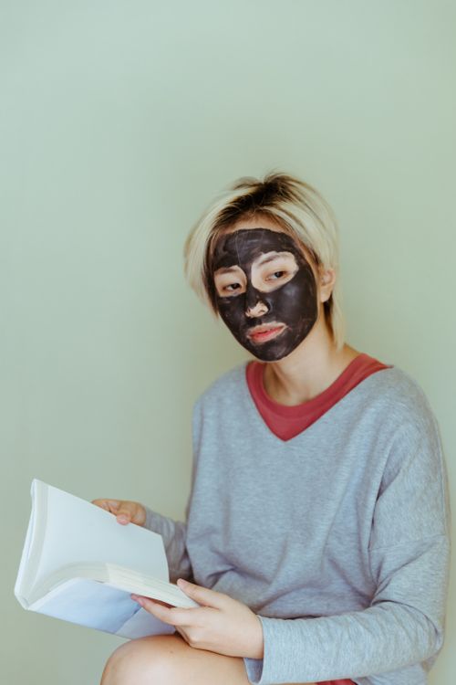 Free Thoughtful young Asian lady in casual clothes with facial mask reading book while sitting on stool against gray wall and looking away pensively Stock Photo