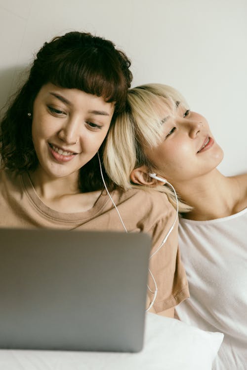 Free Happy young Asian girlfriends in nightwear browsing netbook and smiling while relaxing on floor and listening to music via earphones Stock Photo