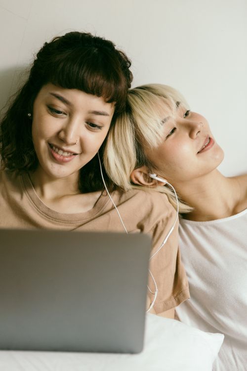 Free Happy young Asian girlfriends in nightwear browsing netbook and smiling while relaxing on floor and listening to music via earphones Stock Photo