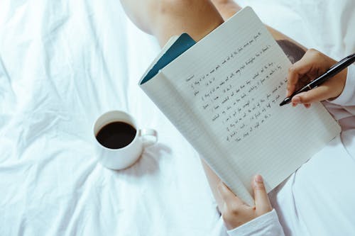 Top view of unrecognizable woman sitting on bed with legs near cup of coffee and writing on notepad with pen while resting at home