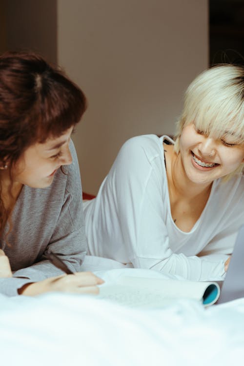 Happy young ethnic women in sleepwear smiling and relaxing in comfortable bed while reading diary and taking notes in modern bedroom