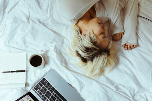 From above ethnic woman lying on bed with laptop coffee and notebook resting while brake in comfortable bedroom at home in weekend