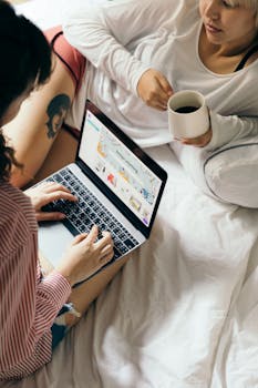 From above of young women using laptop and drinking coffee while resting together on comfortable bed in modern bedroom at daytime