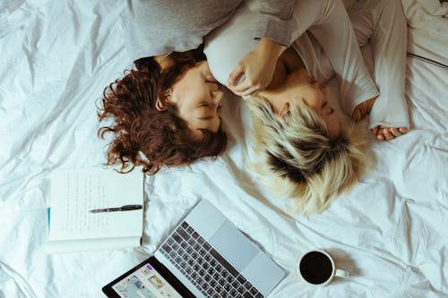 From above of young lesbian couple lying on bed and resting with laptop and cup of coffee while chilling on weekend together