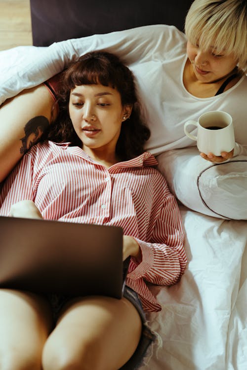 Free From above of young women lying on bed together with laptop while resting and drinking coffee while relaxing during weekend Stock Photo