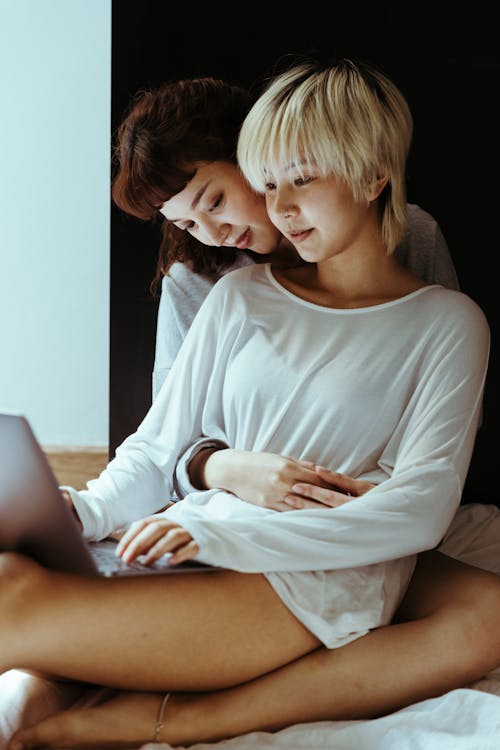 Young ethnic lesbian couple sitting on bed and watching movie on laptop while resting together in comfortable bedroom together during weekend