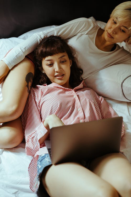 Positive young women lying on bed and browsing laptop while resting in comfortable bedroom together during daytime in morning time