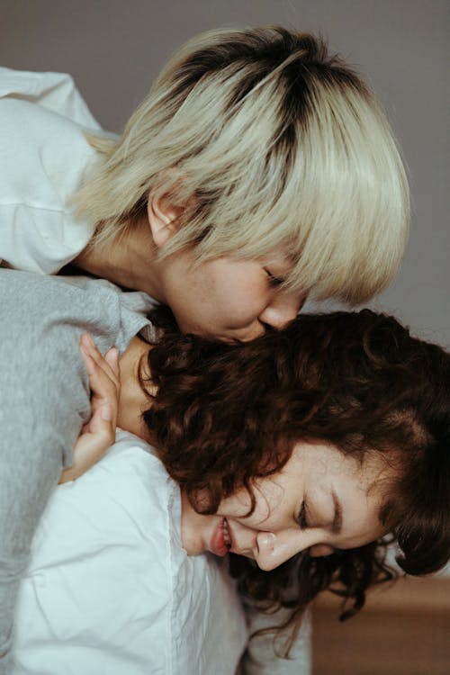 Side view of young ethnic woman hugging and kissing cheerful female while resting on bed in bedroom at home during weekend time and having fun together