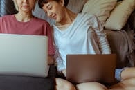 Crop happy Asian girlfriends working on laptops and smiling while sitting together on floor in cozy room with sofa