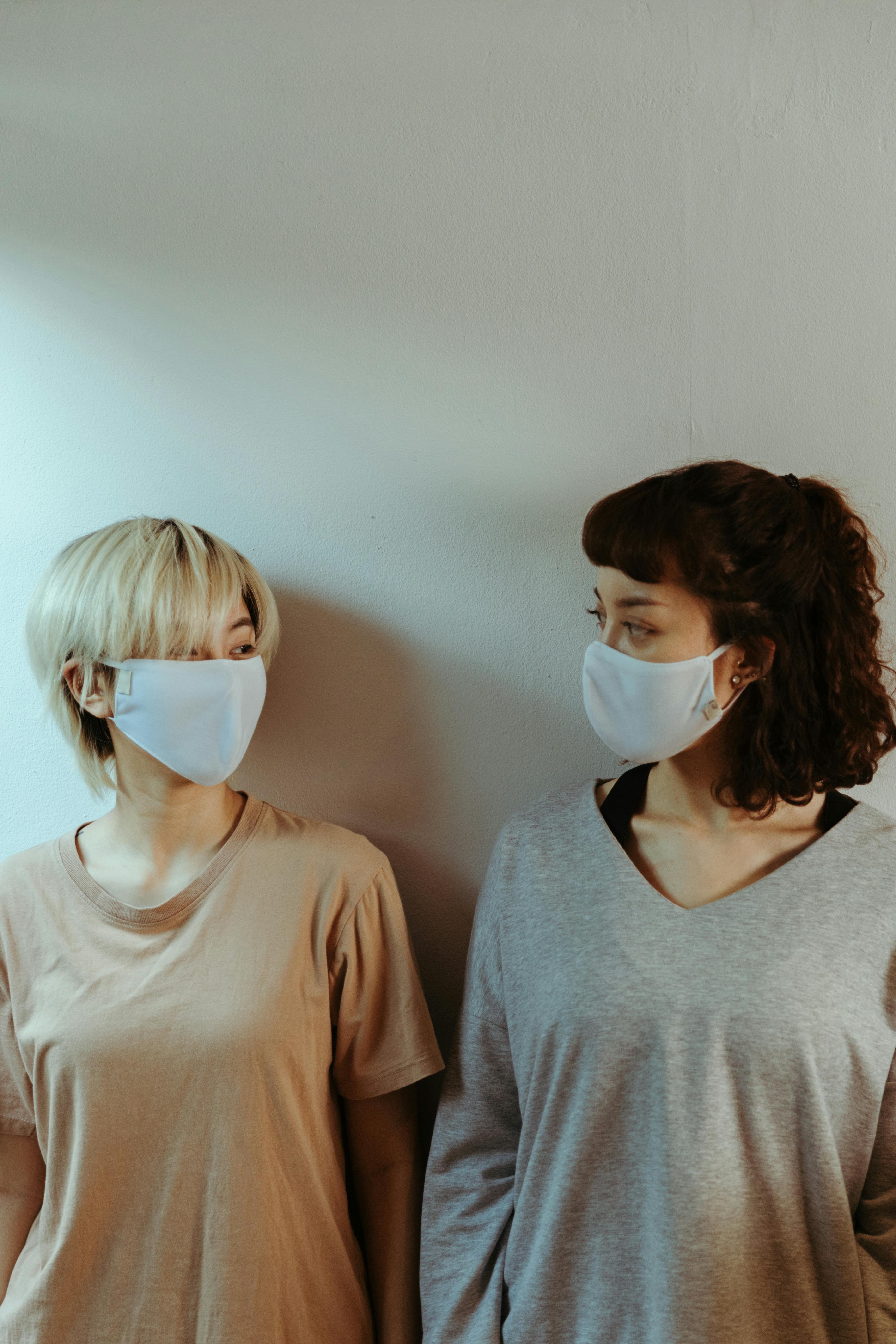 Female friends in white face masks leaning on wall and looking at each other while standing in hall