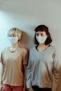 Asian women in home wear in medical masks attentively looking away while standing against white wall