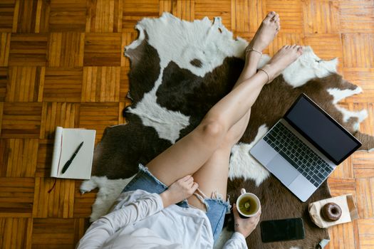 From above of crop woman in jean shorts sitting on floor on cow skin with tea and donut while working remotely on laptop in living room
