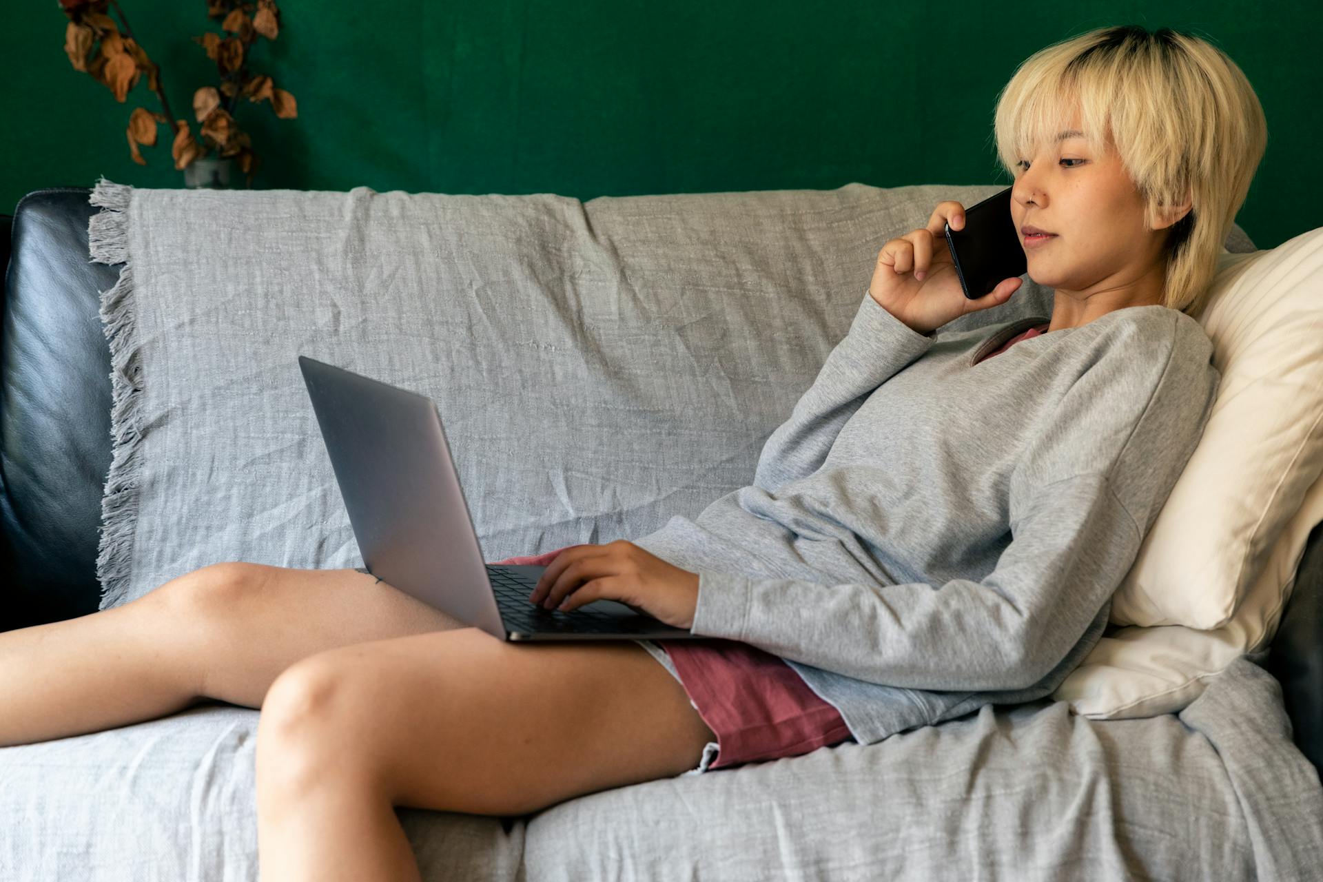 Asian woman working on a laptop and phone while lying on a cozy sofa, remote work concept.