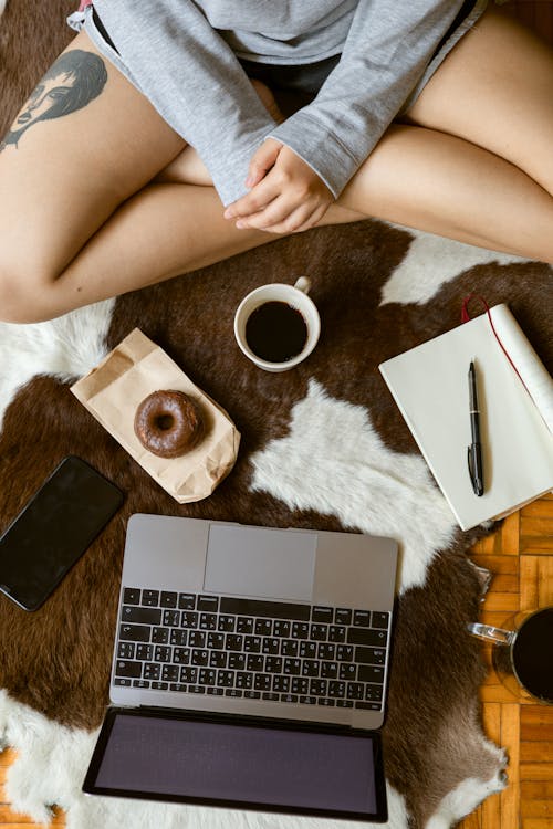 Top view of crop anonymous tattooed female sitting with crossed legs in front of open netbook and cup of coffee with delicious doughnut while studying during breakfast at home