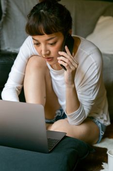 一位穿着休闲装的年轻女性远程工作者坐在公寓床上，一边打电话一边用上网本工作