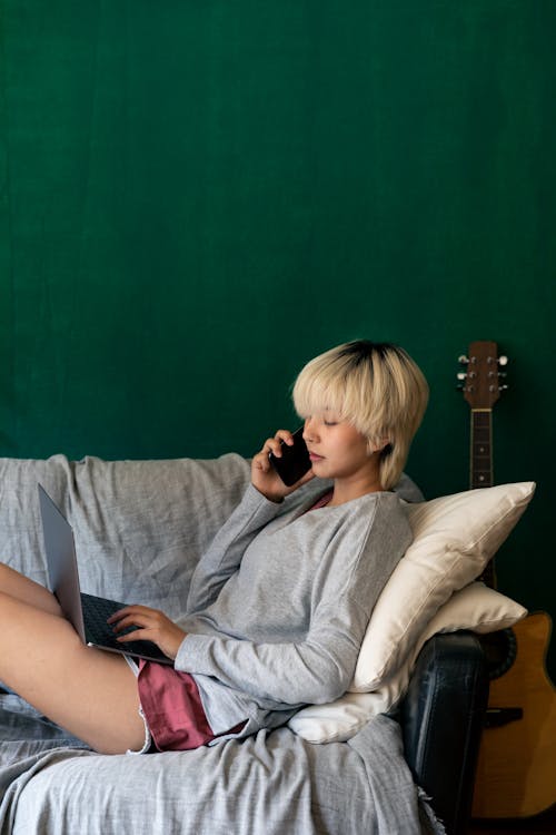 Free Side view of young slender ethnic woman speaking on mobile phone and browsing internet on netbook while lying on comfortable sofa at home Stock Photo