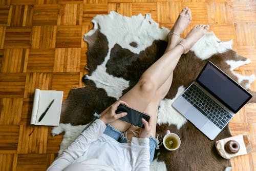 Top view of crop faceless female browsing internet on cellphone while sitting with crossed legs on carpet with tasty doughnut and cup of tea near copybook and laptop at home
