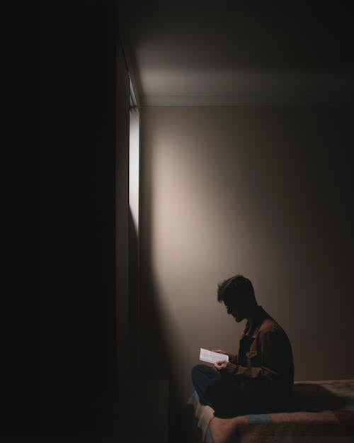 Free A Man Reading Book while Sitting on a Bed Stock Photo