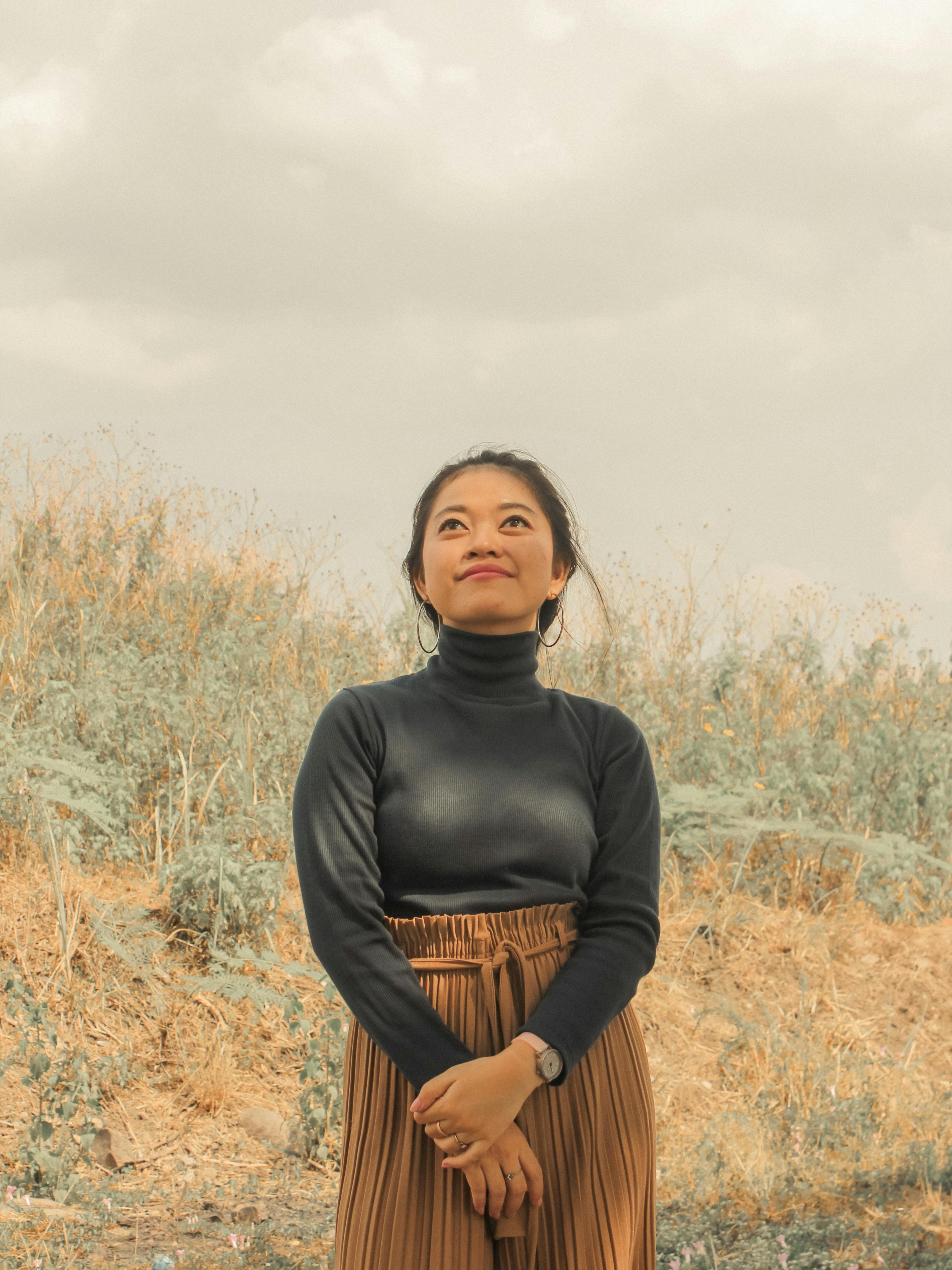 Portrait of a woman in a turtleneck standing in a grassy field, looking thoughtfully at the sky.