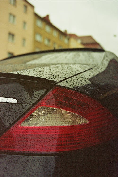 Rear bumper of modern style black car with red taillight and water drops on shiny surface on city street near multistory residential house facade on rainy day