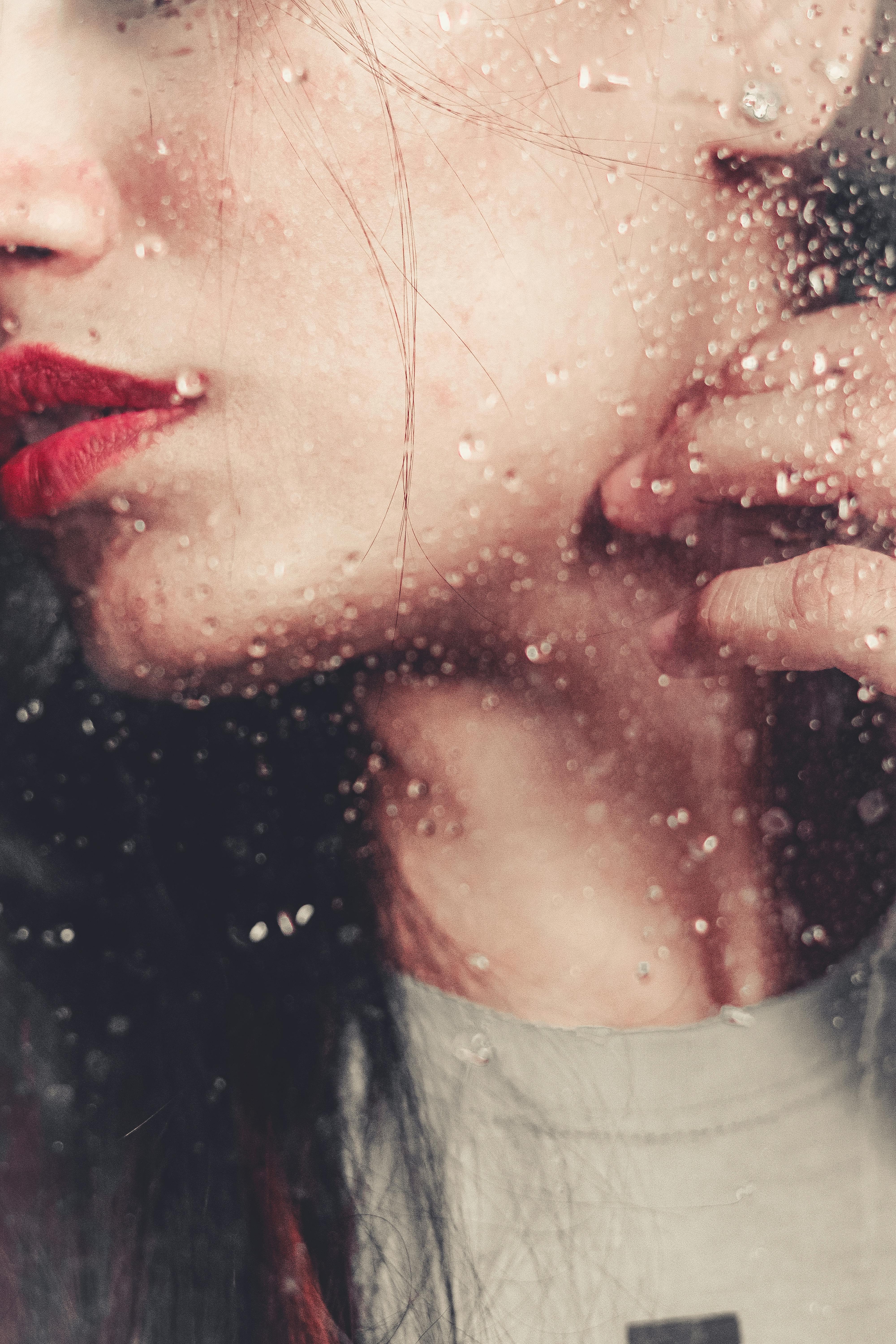 Close-up of woman behind raindrop-covered glass, red lips parted, sensual and contemplative mood
