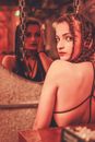 Back view of ethnic female with makeup and pigtails looking over shoulder at camera while standing in front of mirror with different spooky reflection