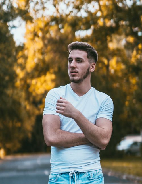 Free Young sporty unshaven male with modern haircut in casual wear contemplating nature while standing on walkway behind bright trees in fall and looking away Stock Photo