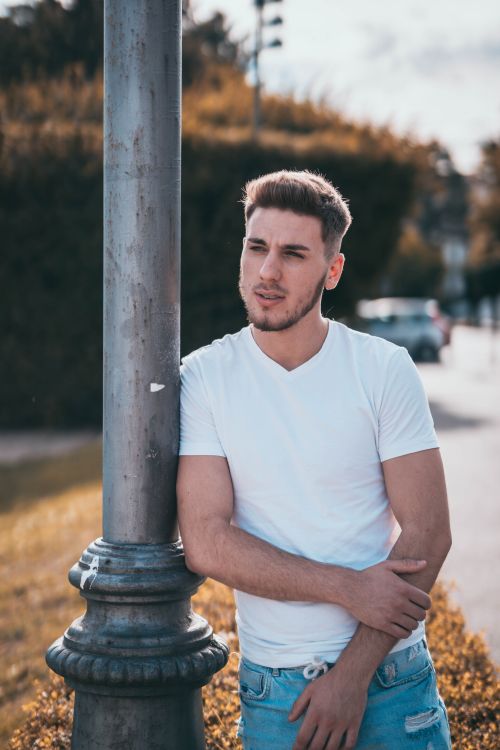 Free Fit young pensive male in jeans and t shirt with contemporary haircut looking away while standing leaned on metal post near asphalt roadway in town in autumn in back lit Stock Photo