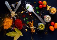 Top view of dry turmeric and paprika near bay leaves with cinnamon sticks and sea salt in spoons near jars with popcorn grains and olives with ripe colorful cherry tomatoes