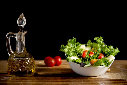 From above of bowl with healthy salad with fresh lettuce leaves and ripe tomato slices on wooden table near glass bottle of olive oil on black background