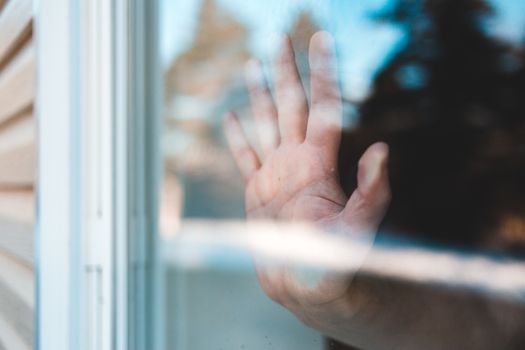 Through glass side view of crop anonymous male touching shiny window with palm of hand standing in apartment in afternoon