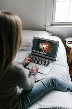 A young woman works remotely on her laptop in a modern, minimalist bedroom setting.