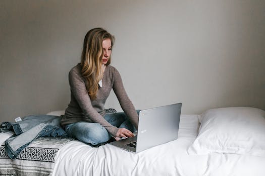 A young woman working remotely on a laptop while sitting on a bed in a cozy, modern bedroom.