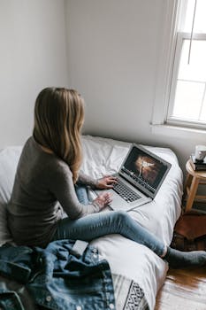 A young woman working from home on a laptop in a cozy, minimalist bedroom setting.
