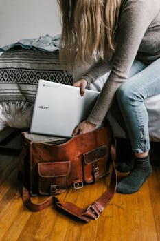 A young woman packs her laptop into a leather bag, preparing for remote work in a cozy bedroom setting.