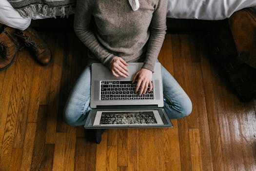 A young woman sitting on a hardwood floor working on a laptop in a modern bedroom setting.