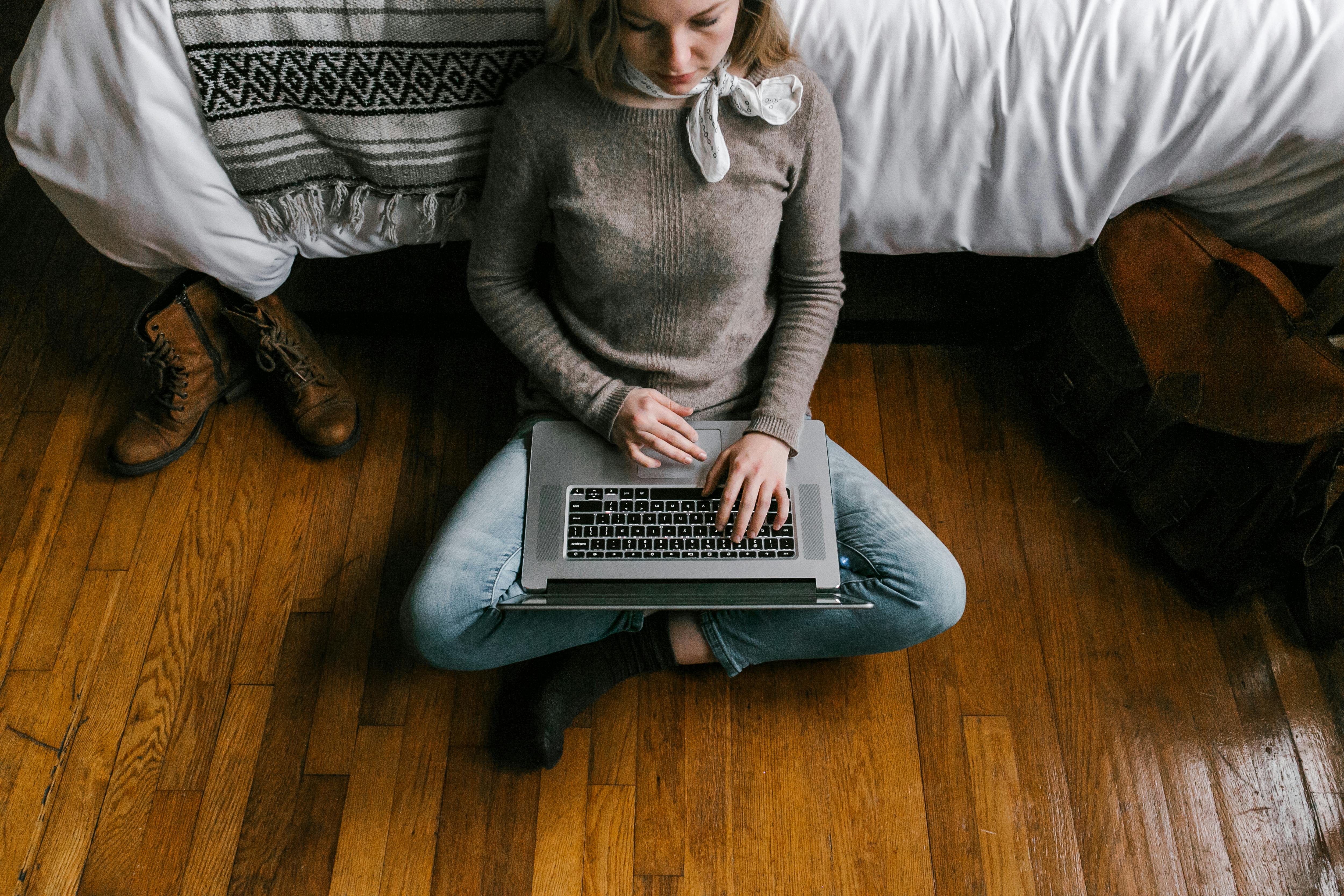 A young woman sitting on hardwood floor working on a laptop in a cozy bedroom setting.