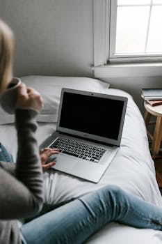 Young woman enjoying coffee while working on laptop in cozy bedroom. Perfect setting for remote work.
