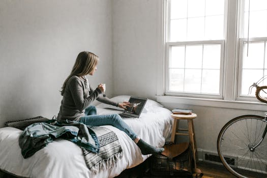 A young woman working from home on her laptop, sipping coffee in a bright bedroom.