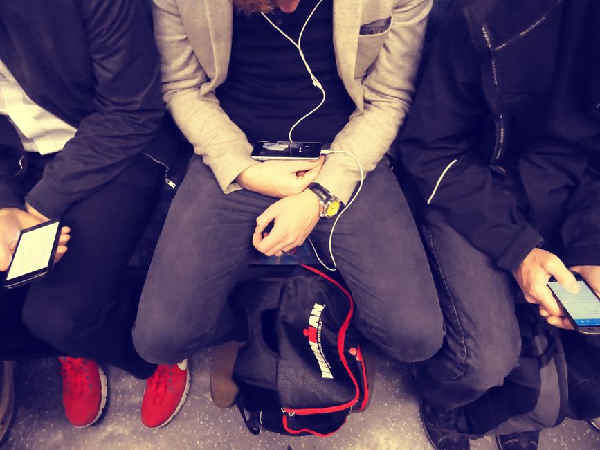 Three men sitting on a subway, using smartphones and earphones, highlighting urban technology use.
