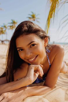 A woman in swimwear smiling while lying on a sunny tropical beach with palm trees.