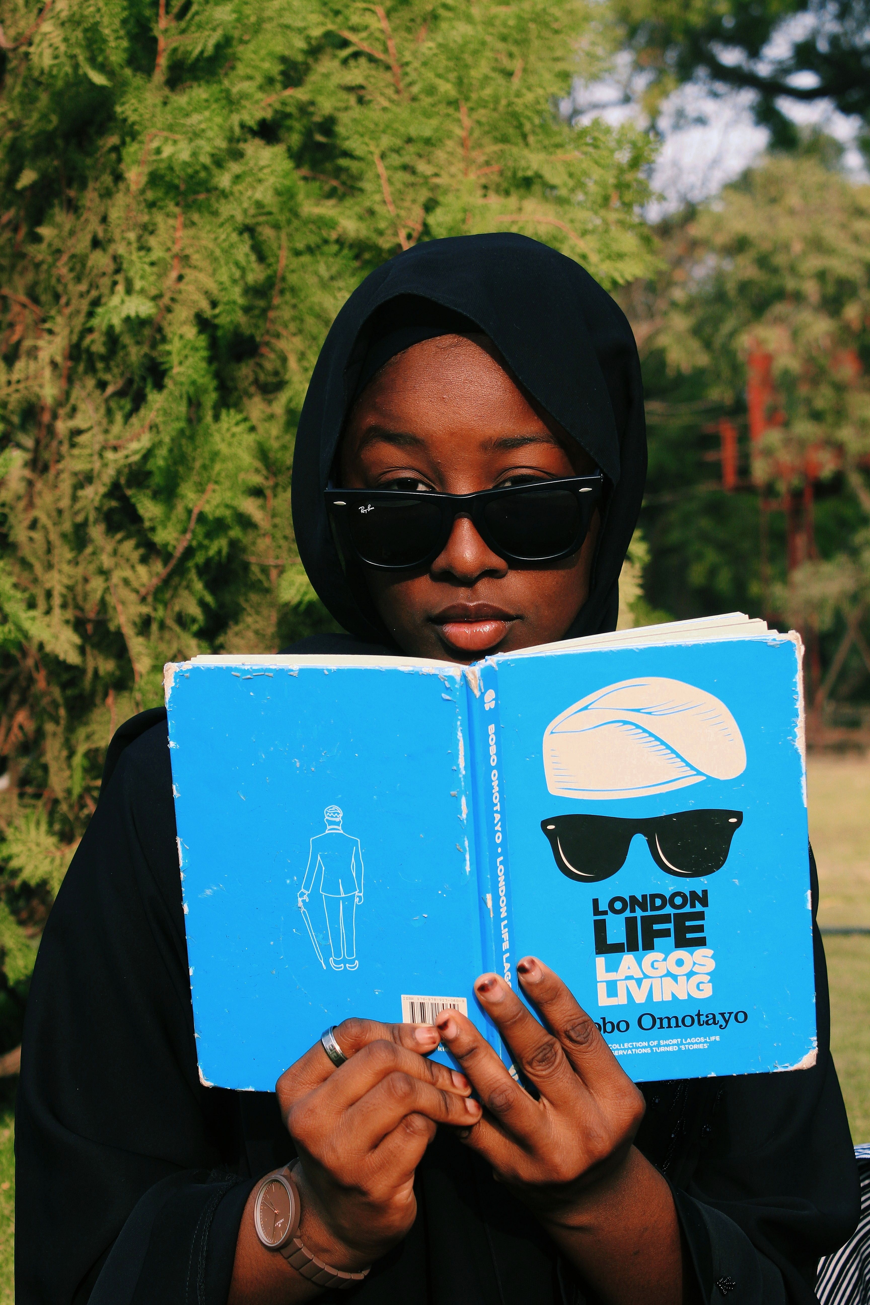 Young focused African American woman in black wear and sunglasses sitting with colorful open book while looking at camera near green trees in garden
